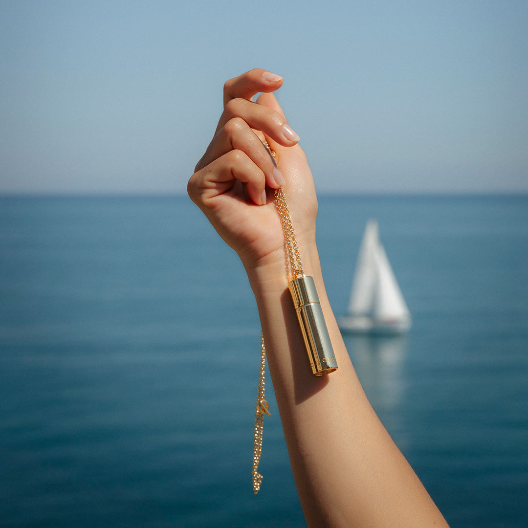 Hand holding Le Spritz necklace in Gold with a scenic background of the ocean and a sailboat.